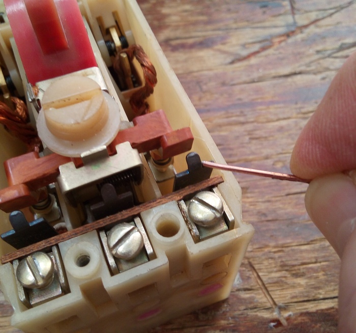 Проволочкой, здесь указана биметаллическая пластина.Нагреваясь от подходящего через неё большого тока, она постепенно изогнётся и нажав на деталь из пластика, освободит защелку. Реле сработает.